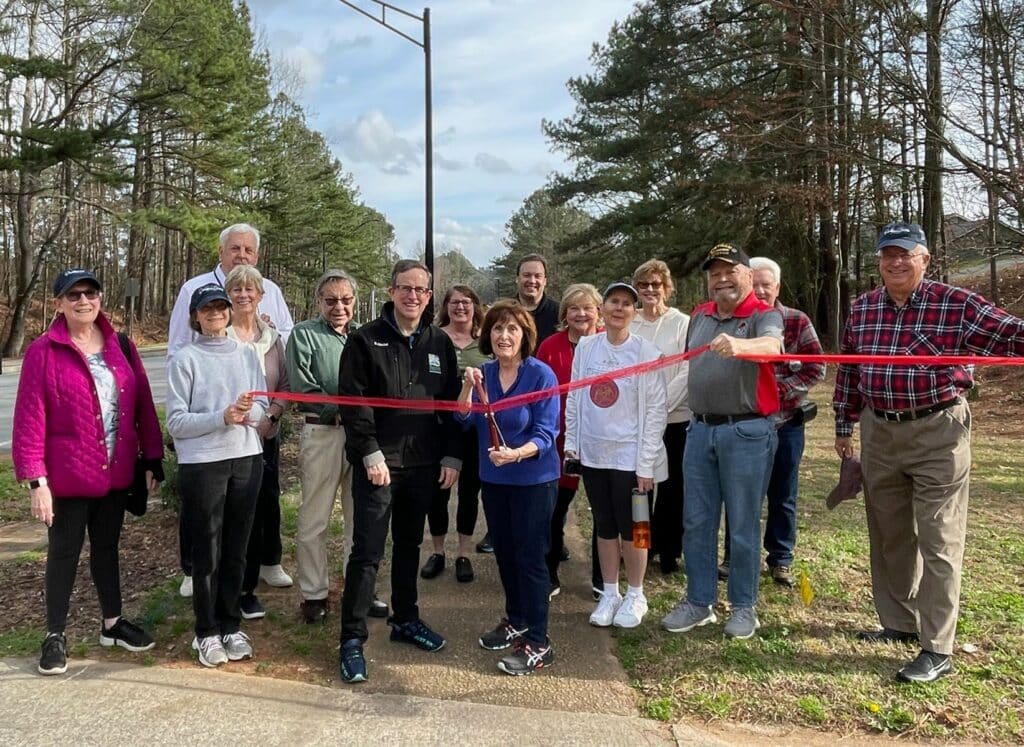 Susan Albright (with scissors) and Peachtree Corners Councilmember Eric Christ (in black jacket), along with Dunwoody Manor neighbors, ready to cut the ribbon with one of the new street lights in the background. 