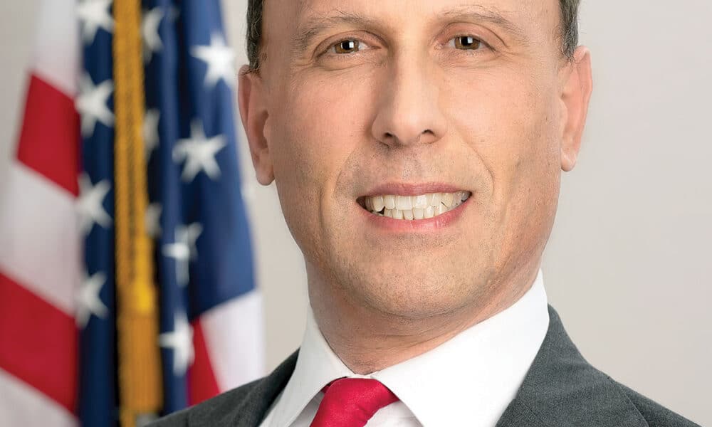 Headshot of a smiling white, male city politician in a suit and tie standing in front of an American flag.