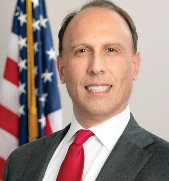 Headshot of a smiling white, male city politician in a suit and tie standing in front of an American flag.