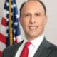 Headshot of a smiling white, male city politician in a suit and tie standing in front of an American flag.