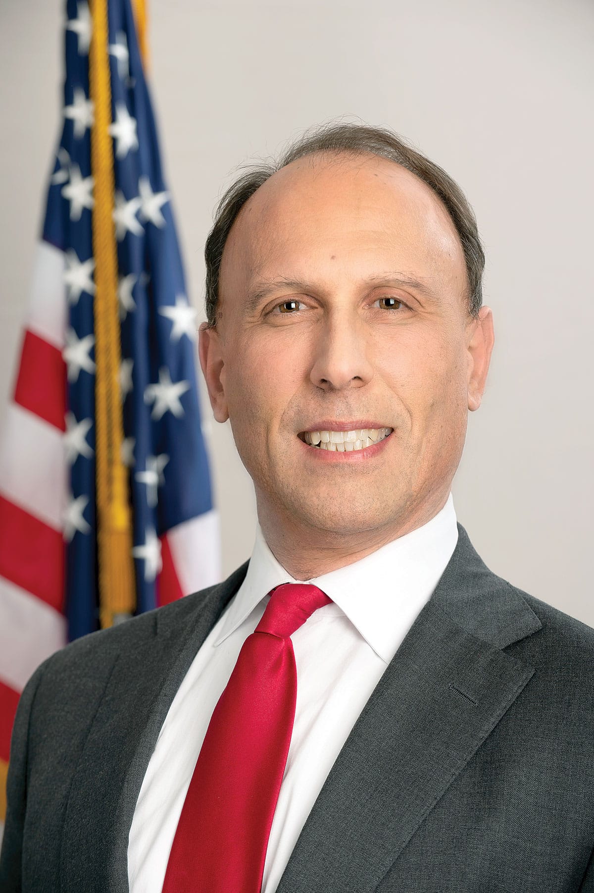 Headshot of a smiling white, male city politician in a suit and tie standing in front of an American flag.