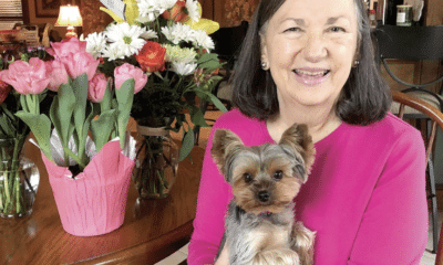 A woman in a pink sweater holding a small dog and sitting at a table with pink and white flowers