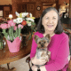 A woman in a pink sweater holding a small dog and sitting at a table with pink and white flowers