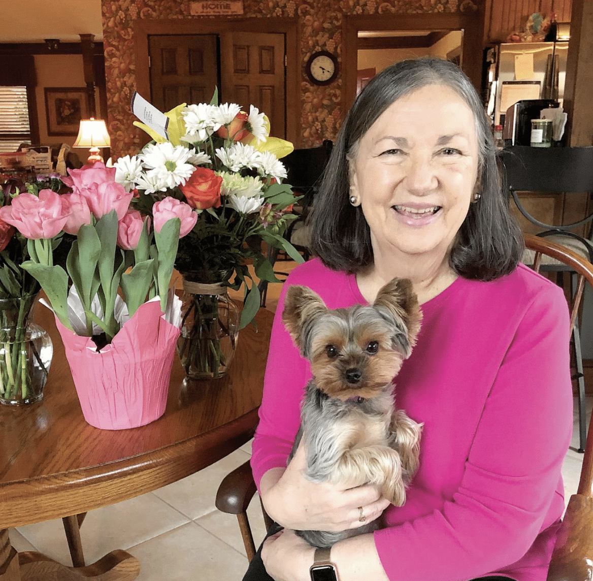 A woman in a pink sweater holding a small dog and sitting at a table with pink and white flowers