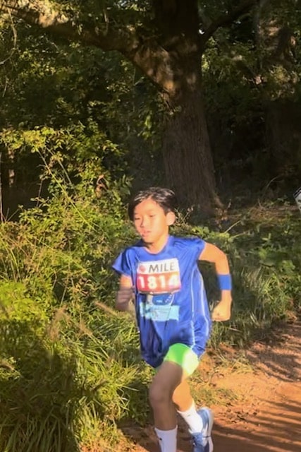 Young cross country runner in a blue shirt running on a dirt trail in a wooded area.