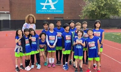 A group of young cross country track students in blue shirts standing with their coach in front of a school building.