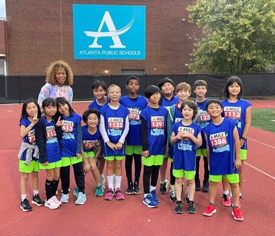 A group of young cross country track students in blue shirts standing with their coach in front of a school building.