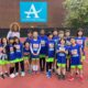 A group of young cross country track students in blue shirts standing with their coach in front of a school building.