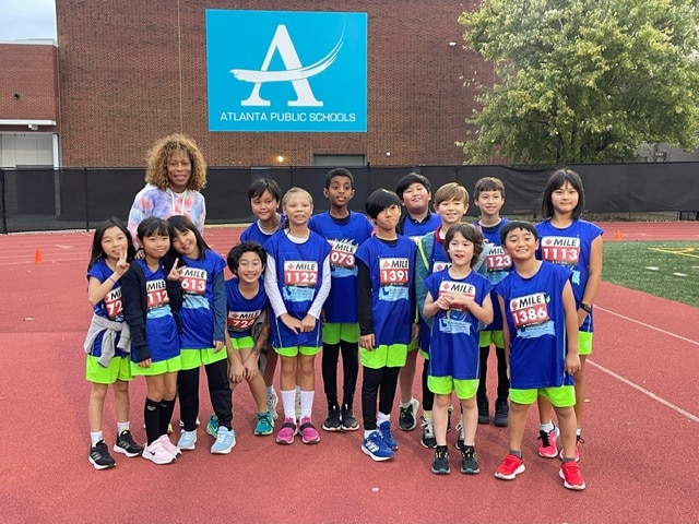 A group of young cross country track students in blue shirts standing with their coach in front of a school building.