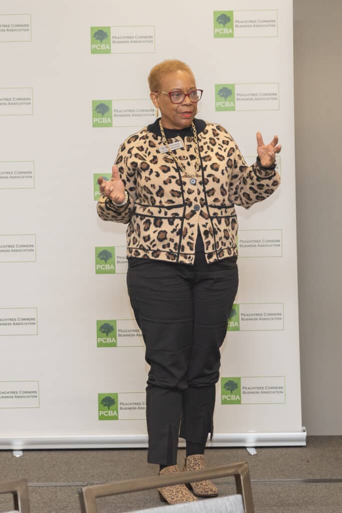 A short-haired woman in black pants and print top standing in front of a PCBA event backdrop