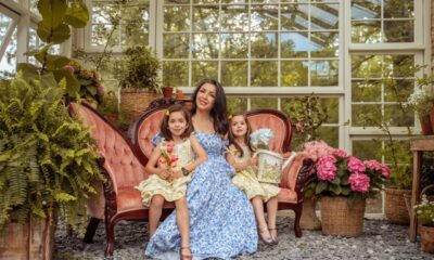 A woman with long dark hair wearing a blue dress, sitting on a pink Victorian sofa with two young girls in a greenhouse space