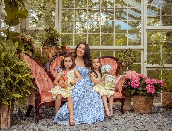 A woman with long dark hair wearing a blue dress, sitting on a pink Victorian sofa with two young girls in a greenhouse space