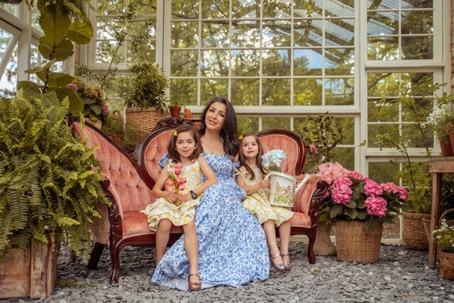 A woman with long dark hair wearing a blue dress, sitting on a pink Victorian sofa with two young girls in a greenhouse space