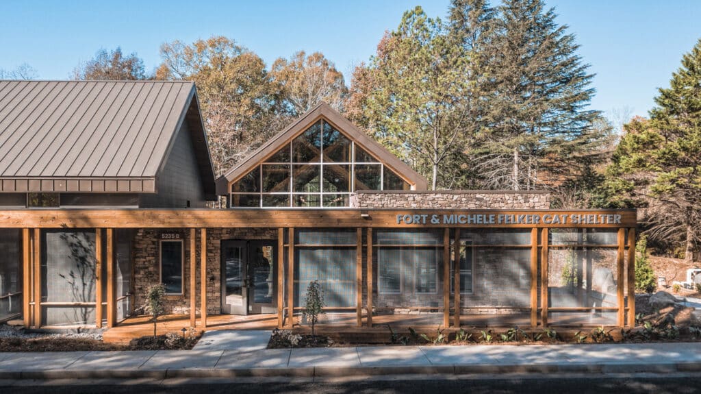 Wood framed building with lots of windows and a screened-in patio set within a lightly wooded lanscape.