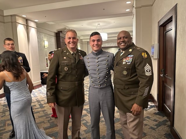 A young man in military school uniform standing in a hallway between two Army men All of them are smiling for the camera.