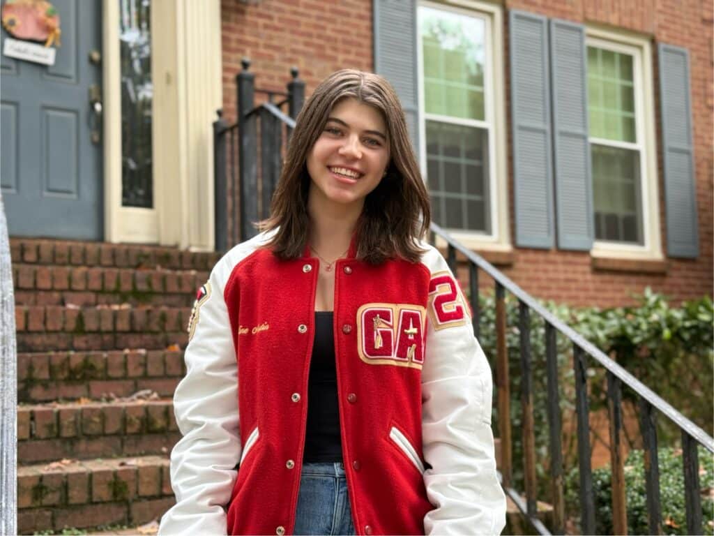 Female high school student with shoulder-length brown hair standing in front of a building with steps, wearing a red and white school sports jacket