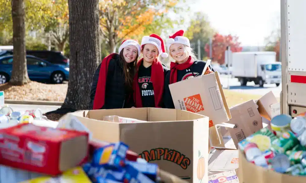 Three female students wearing santa hats and red scarves, posing in front of large boxes of donated food items they've helped gather for the Salvation Army can-a-thon.