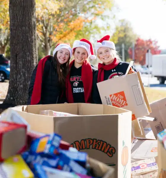 Three female students wearing santa hats and red scarves, posing in front of large boxes of donated food items they've helped gather for the Salvation Army can-a-thon.