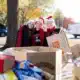 Three female students wearing santa hats and red scarves, posing in front of large boxes of donated food items they've helped gather for the Salvation Army can-a-thon.