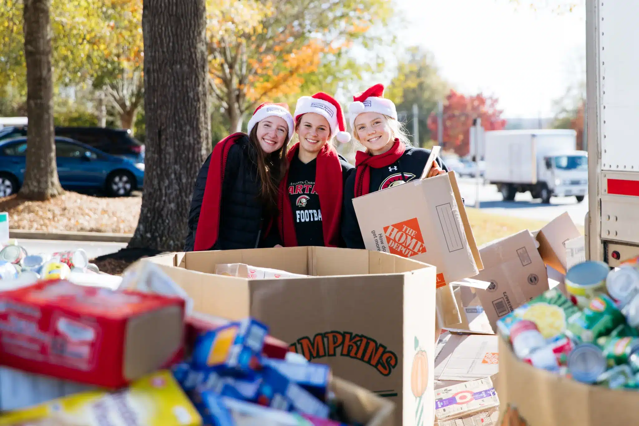 Three female students wearing santa hats and red scarves, posing in front of large boxes of donated food items they've helped gather for the Salvation Army can-a-thon.