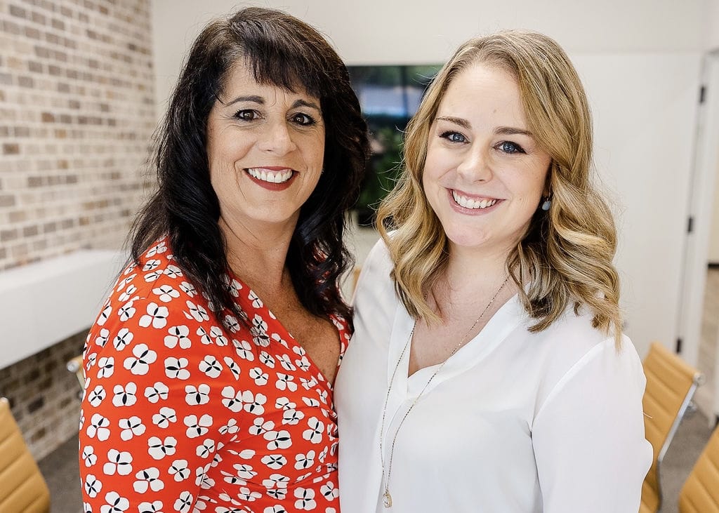 A dark-haired woman wearing a red print blouse standing next to her blonde-haired adult daughter who is wearing a white blouse