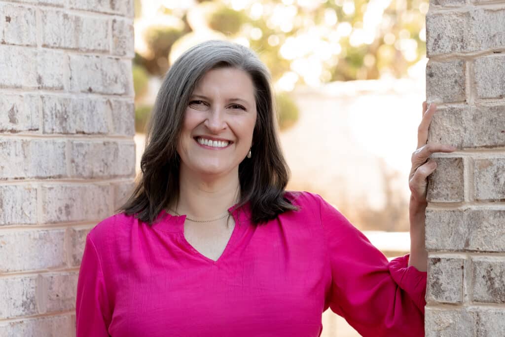 Middle-aged woman with shoulder length, brown/grey hair wearing a pink long-sleeved blouse and smiling at the camera