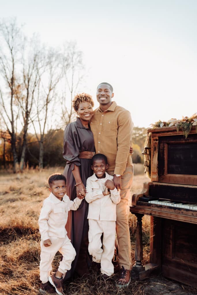 Beautiful Black family with mom and dad and two young sons posing outdoors next to a brown piano in fall