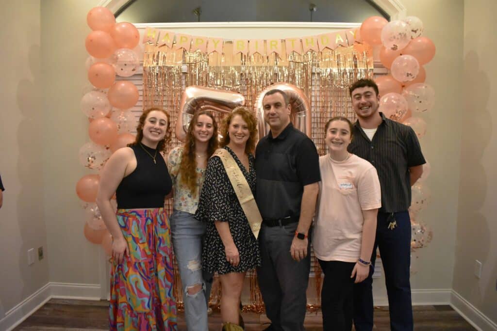 Family of six — mom, dad, and four teen and adult kids standing in front of holiday decorations and pink balloons 