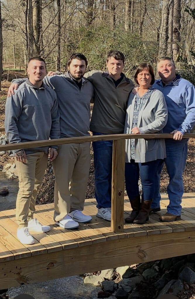 Middle aged mom and dad with their three grown sons standing on a short wooden bridge in a wooded area