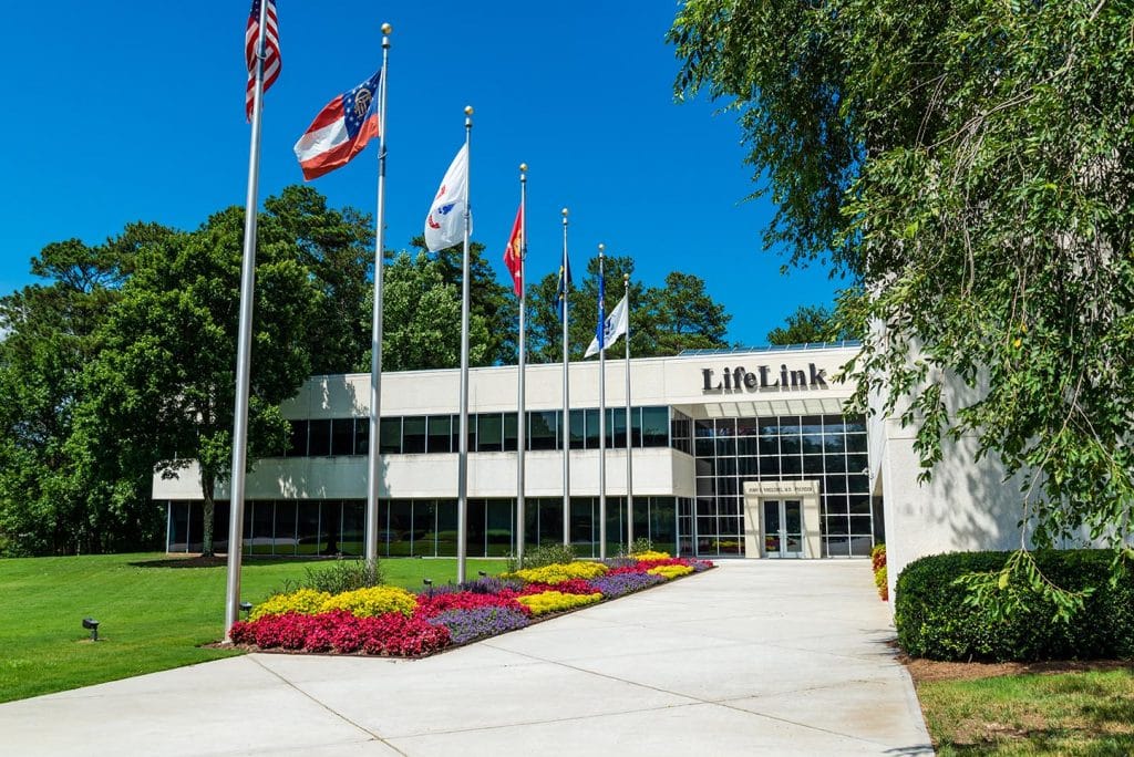 Mid-sized white and class building with wide walkway leading up to it. The walkway is lined with flags and there are trees and colorful plants.