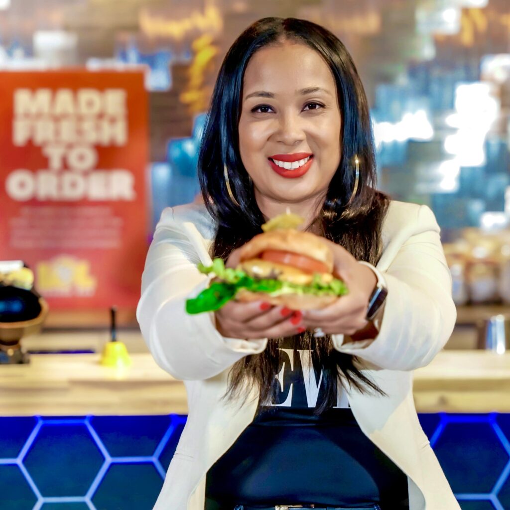 Smiling woman with long dark hair, holding a burger in front of her as if to give it to someone.