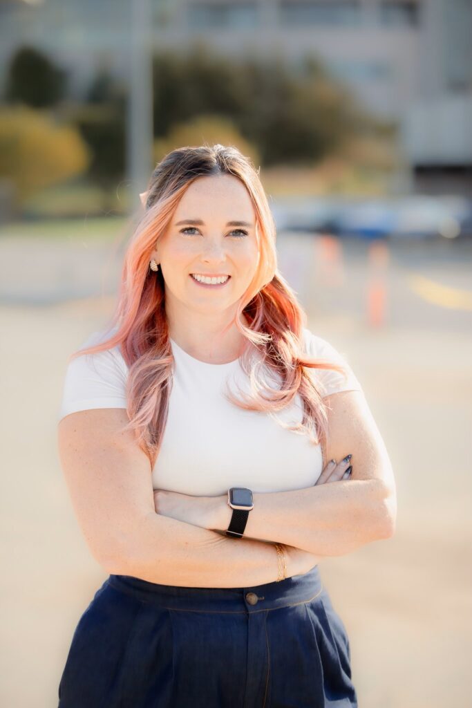 Young woman with long pink and brown hair, wearing a white top and dark blue pants. She's standing outside with her arms crossed in front of her.