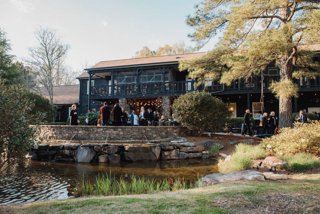 Exterior of the main building of Furkids surrounded by woods, greenspace and a small water feature. People are gathered on the patio for a party.