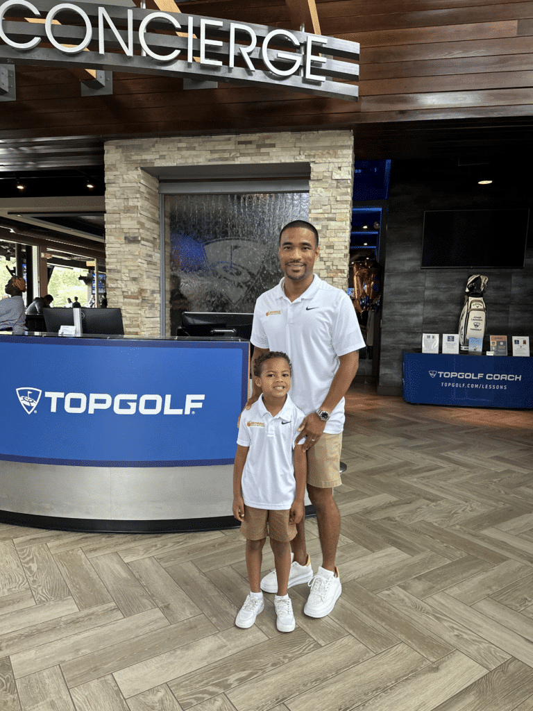 A Black father with his young son at a Top Golf location. They are both wearing white shirts and tan shorts, standing the lobby for  the photo.