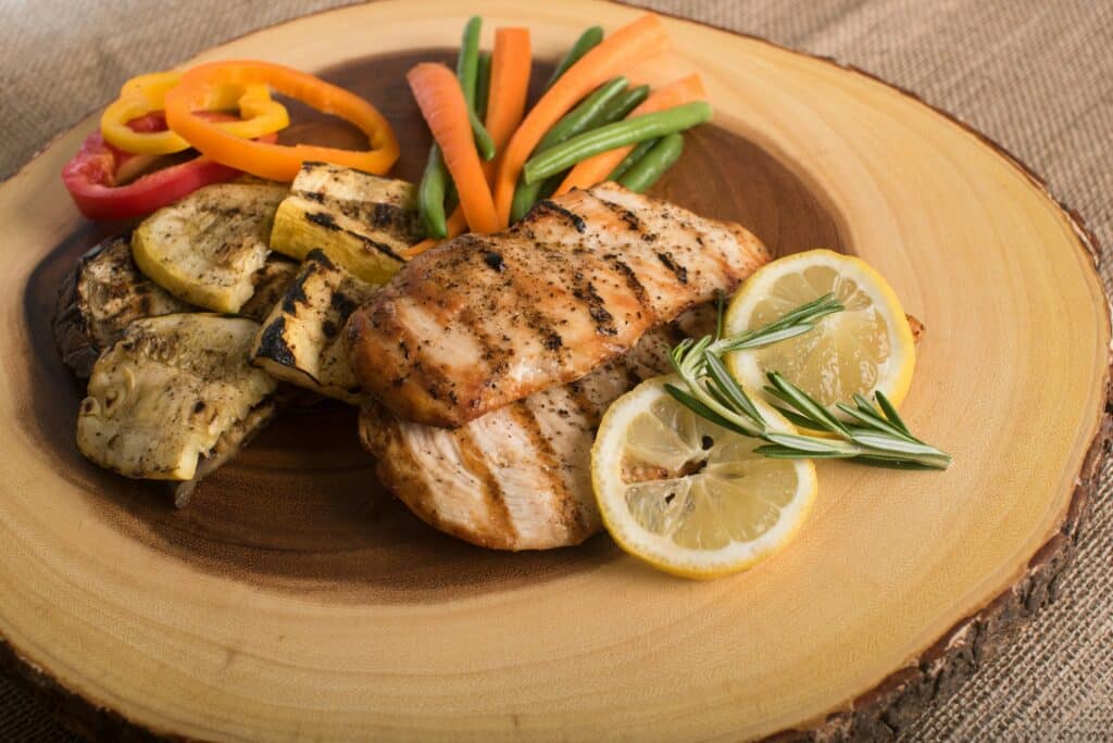 Grilled chicken breast with vegetables and lemon slices plated on a round piece of wood