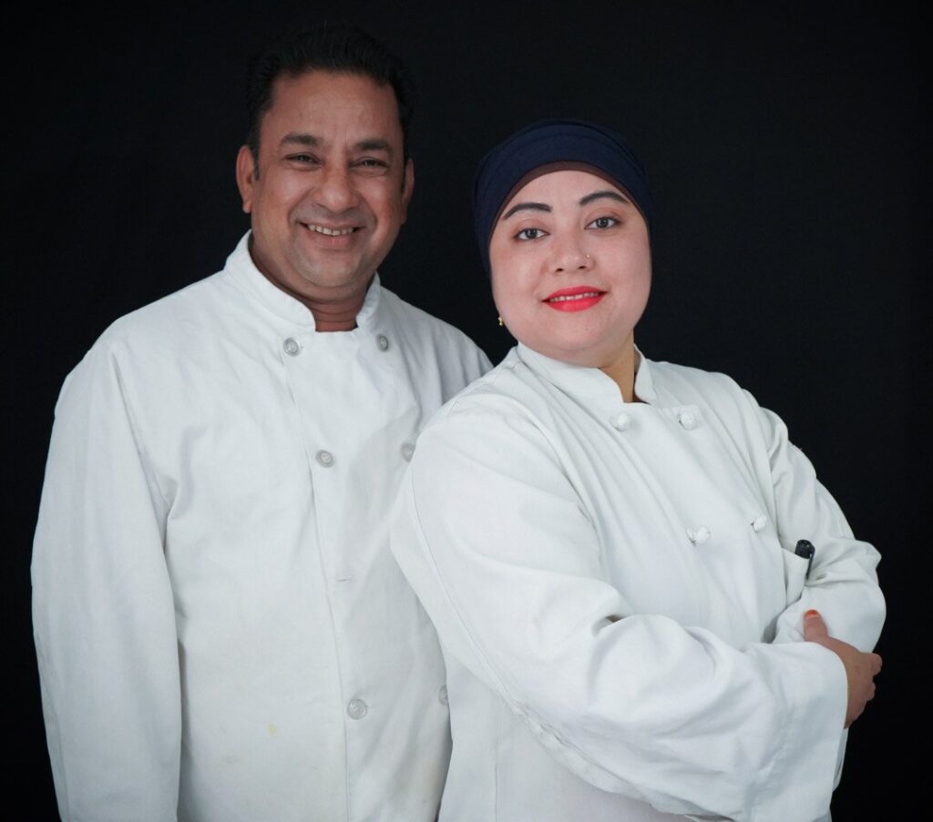 A male chef and female chef wearing white chef coats standing in front of a black background