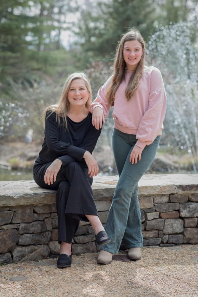 A mom and daughter, both with long blonde/light brown hair. Mom is wearing a black top and pants, sitting on a low stone wall; daughter is standing beside her, wearing a pink top and light blue jeans.