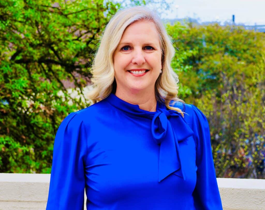 Middle aged woman with shoulder-length blonde hair, wearing a bright blue top, standing outside in front of greenery.