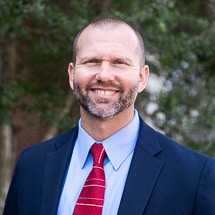 White man in a light blue shirt, dark blue blazer and red tie. He has very short hair and a short mustache and beard.