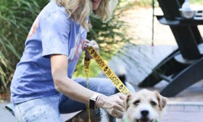 Blonde haired woman wearing sunglasses, kneeling down to pet a brown and white dog on a leash. They are outside on a patio with green plants behind them.