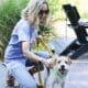 Blonde haired woman wearing sunglasses, kneeling down to pet a brown and white dog on a leash. They are outside on a patio with green plants behind them.