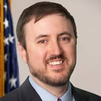 White man with short brown hair and facial hair wearing a suit, smiling at the camera with an American flag behind him.