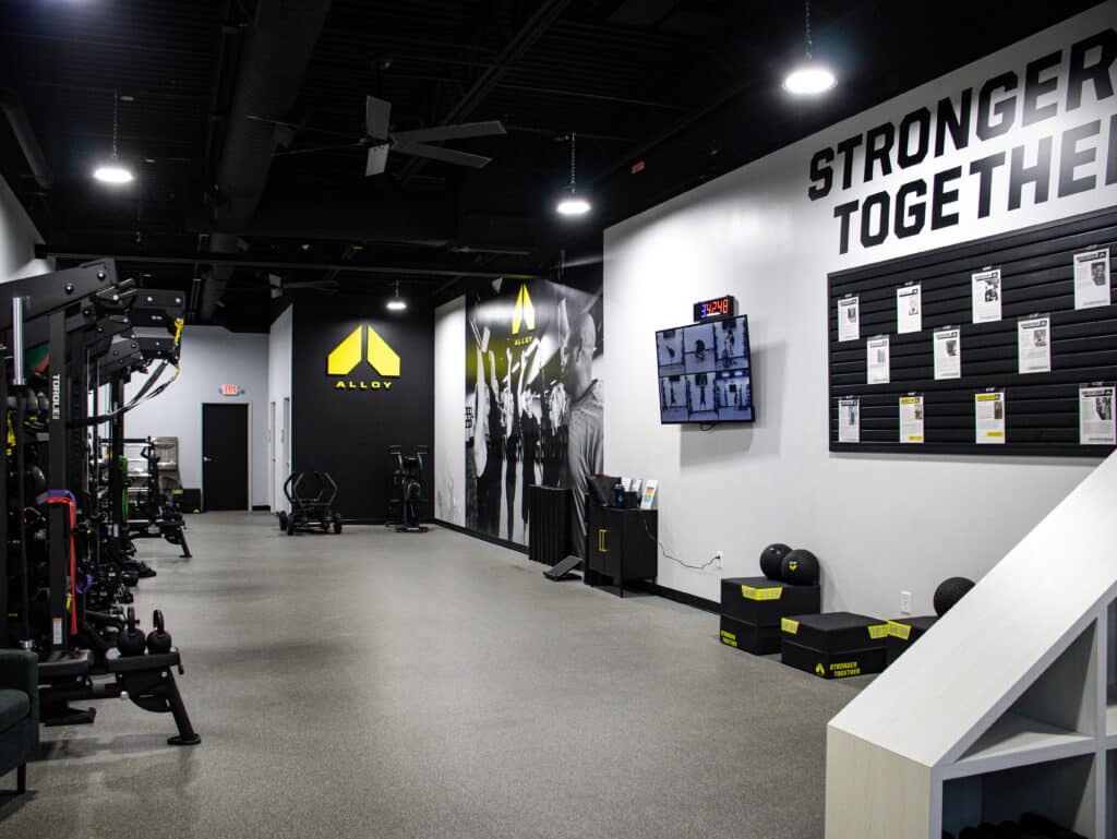 Interior of Alloy Personal Training studio. The walls are white with a black painted ceiling. Stronger Together is painted in black on the right side wall. Fitness equipment lines the left side of the room. The floor is grey and everything is organized and tidy.