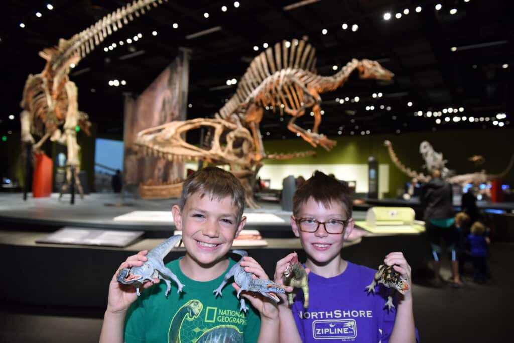 Two smiling, young boys holding toy dinosaurs at a dinosaur exhibit at a natural history museum