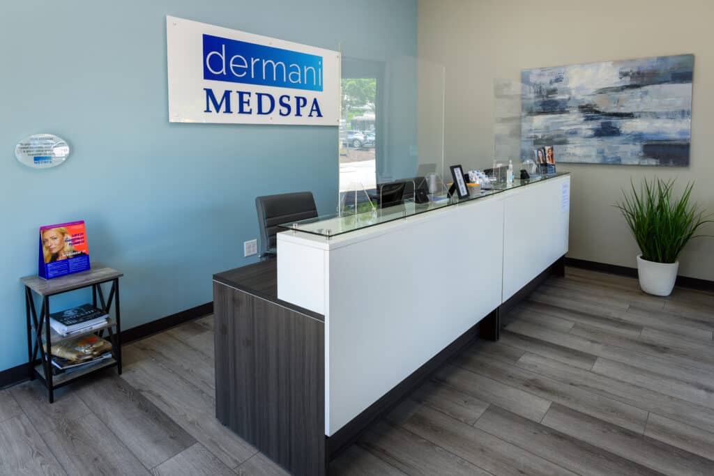 Front desk area of the Dermani MedSpa in Peachtree Corners. The reception desk is long and white, the wall colors are light blue and cream and there's a painting on one wall above a potted plant in a white pot.