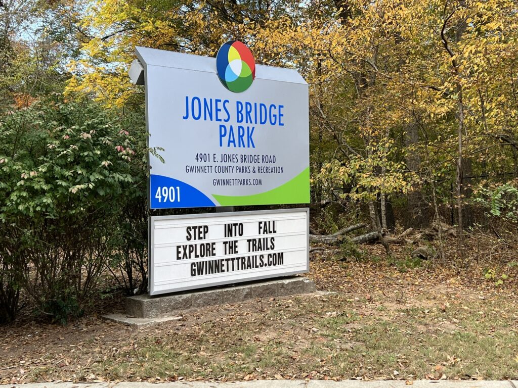 Entrance sign to Jones Bridge Park in Gwinnett County, set against a small wooded area with fall trees.