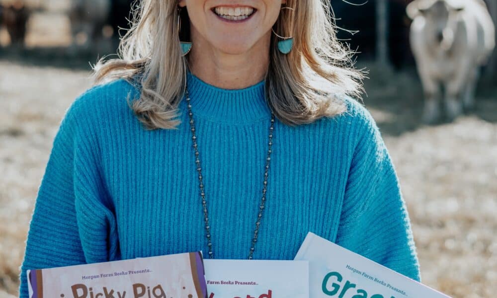 Middle-aged woman with blonde hair, smiling and wearing a blue shirt. She's standing with a building and a cow behind her, and she's holding copies of the three children's books she's written.