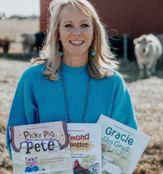 Middle-aged woman with blonde hair, smiling and wearing a blue shirt. She's standing with a building and a cow behind her, and she's holding copies of the three children's books she's written.