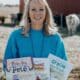 Middle-aged woman with blonde hair, smiling and wearing a blue shirt. She's standing with a building and a cow behind her, and she's holding copies of the three children's books she's written.