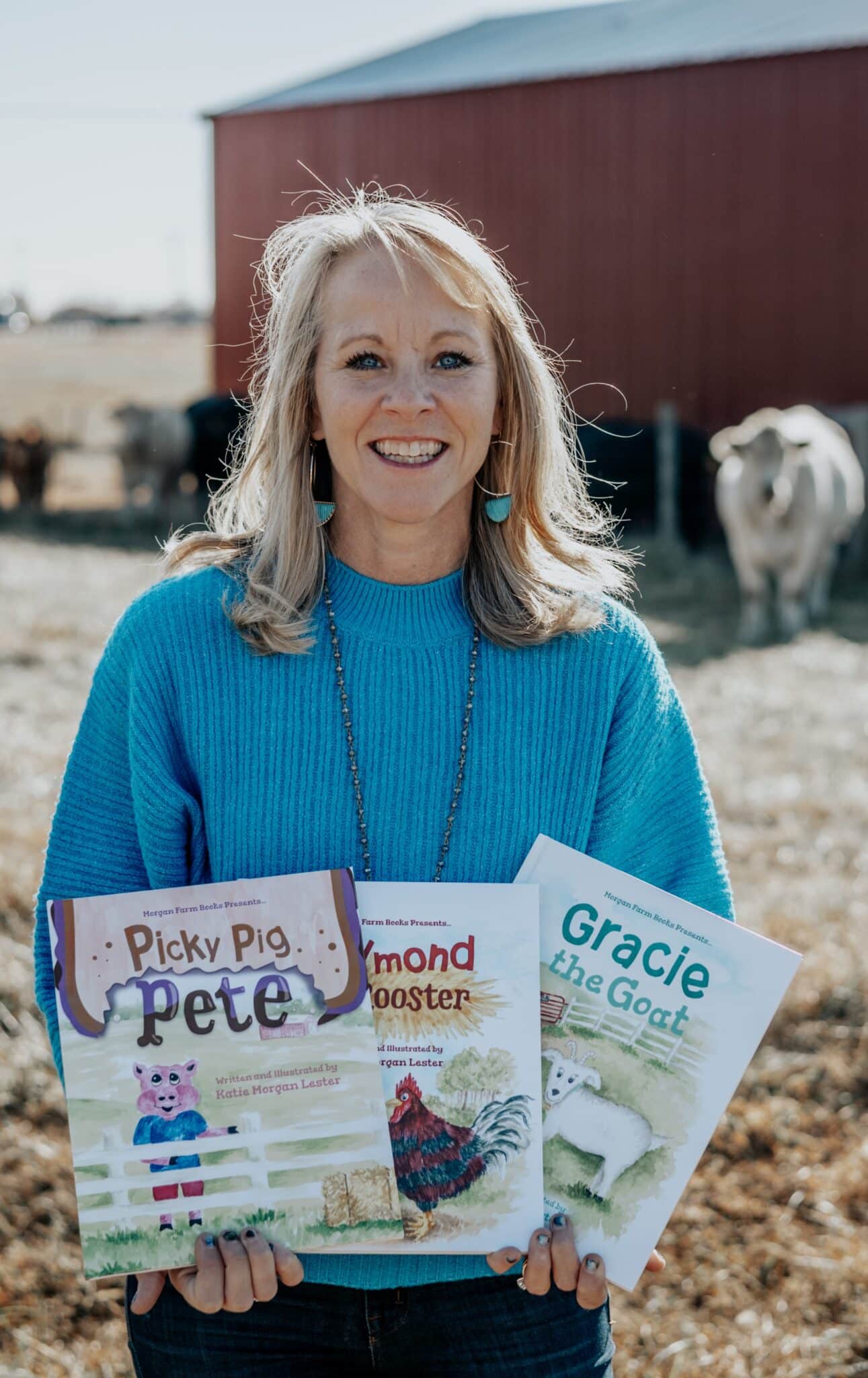 Middle-aged woman with blonde hair, smiling and wearing a blue shirt. She's standing with a building and a cow behind her, and she's holding copies of the three children's books she's written.